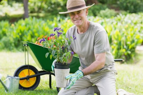 A smiling older man wearing a straw hat, light grey t-shirt, and gardening gloves kneels on a well-maintained grassy lawn in a landscaped outdoor garden in Erith, Kent, holding a potted flowering plant with purple and red blooms. Behind him, a green wheelbarrow with large yellow-and-black wheels is positioned on the lawn, filled with gardening tools and additional plants. The garden features a neatly trimmed hedge along the background, with a mix of trees and shrubs providing a natural border, and a stone-paved pathway visible to the right. The scene is illuminated by soft natural light, suggesting a clear, mild day. The overall setting reflects a tidy, herbaceous outdoor space suitable for gardening services such as lawn care, planting, and landscape maintenance offered by Lawn Mowing Erith, with a focus on natural textures, vibrant plant colours, and well-kept greenery typical of a professional gardening environment in the Erith area.